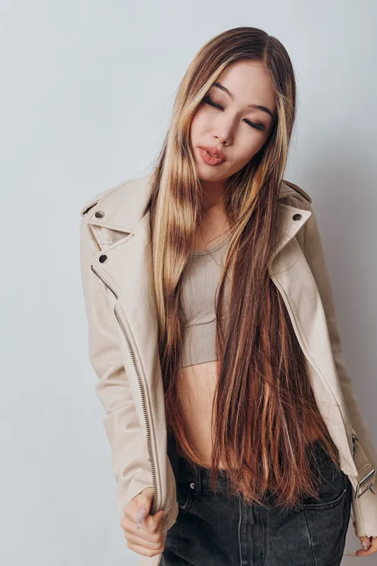 Young woman with long hair wearing beige jacket and crop top posing on light background for fashion style portrait and casual beauty look indoors.