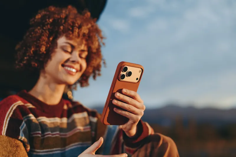Woman smiling outdoors, curly hair in a soft breeze, holding smartphone for a casual selfie, warm sunset light, relaxed lifestyle portrait, scenic hills background, natural colors