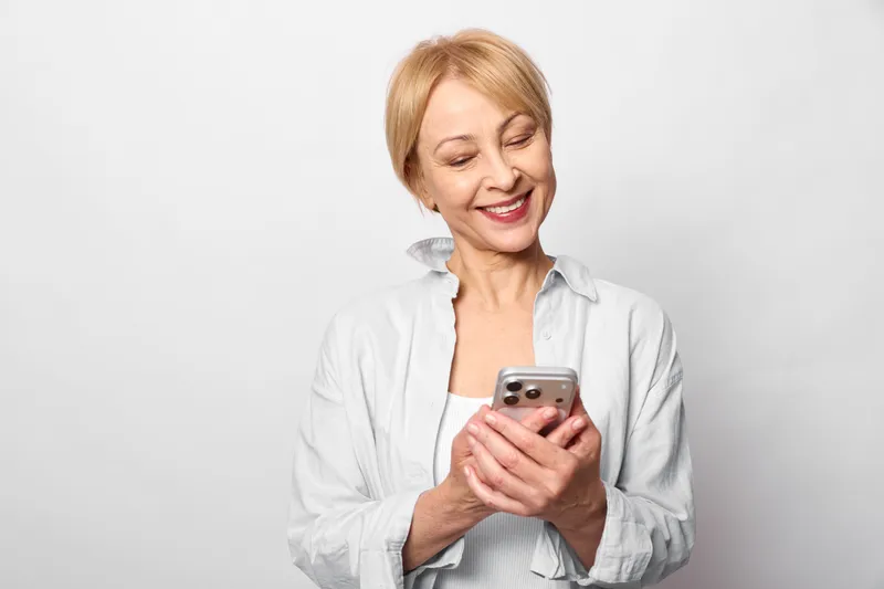 Cheerful senior woman holding smartphone, smiling at a message, studio portrait on white background, relaxed hands, calm expression, concept modern communication