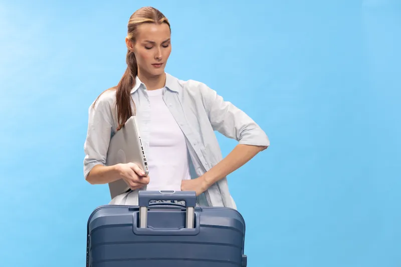 Young woman in casual clothes packing a blue suitcase with a laptop on a blue background. Concept of travel preparation, business trip, and vacation planning.