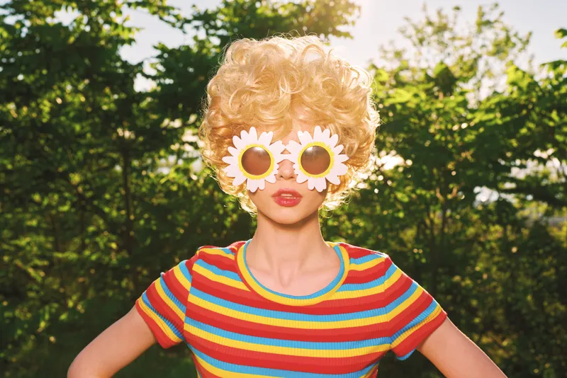 A playful woman outdoors wears a bright striped shirt and flower sunglasses, embracing a retro lifestyle. Sunny light and bold colors convey whimsical fashion and creative self expression.