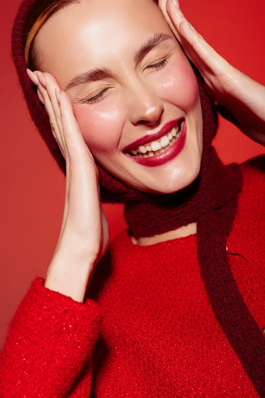 Smiling woman in red sweater and scarf touches her face with closed eyes. Bright red background and warm lighting create a joyful and cozy winter fashion concept.