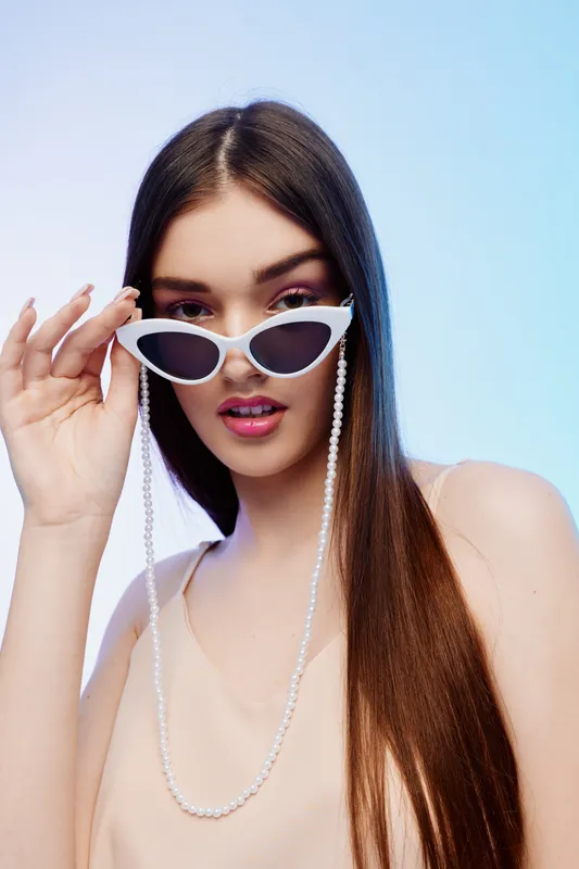 Young woman beauty fashion model with long straight hair wearing white cat eye sunglasses and a pearl chain. Studio portrait on a pastel gradient background. Concept for style and elegance.