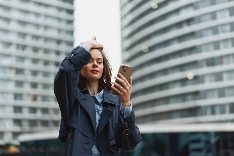 Young woman with long brown hair wearing a dark coat uses a smartphone outdoors in an urban environment during the daytime.