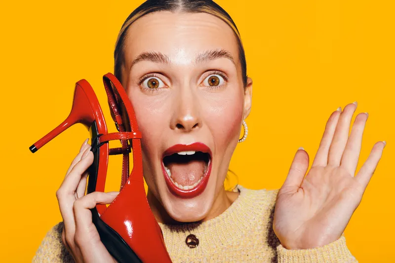 Surprised woman with red lipstick holds red high heel shoe near face on bright yellow background. Concept of fashion, style, excitement, and vibrant emotions in studio lighting.