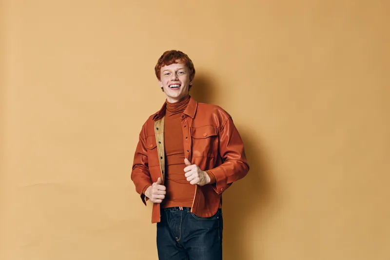 Young man with curly hair smiling while wearing orange jacket and sweater against beige background. Casual style and happiness with confident posture and jeans.