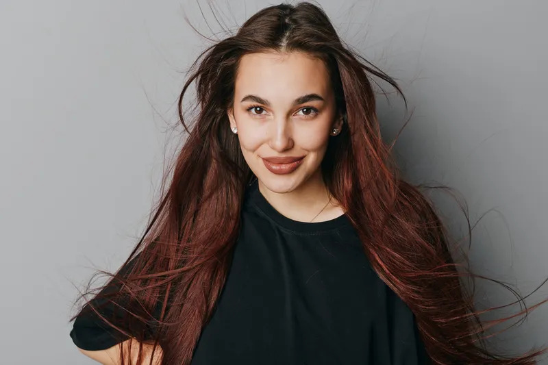 Young woman with long hair smiling confidently at camera wearing black shirt against neutral background. Portrait of female model with natural makeup and casual style indoors.