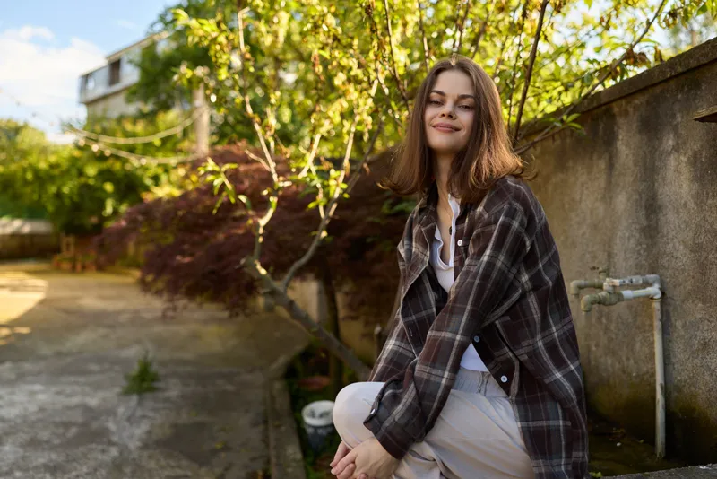 A young woman enjoys a casual outdoor lifestyle moment in daylight, wearing a plaid jacket and light dress, leaning against a wall with a serene smile. Concept: everyday fashion and relaxed
