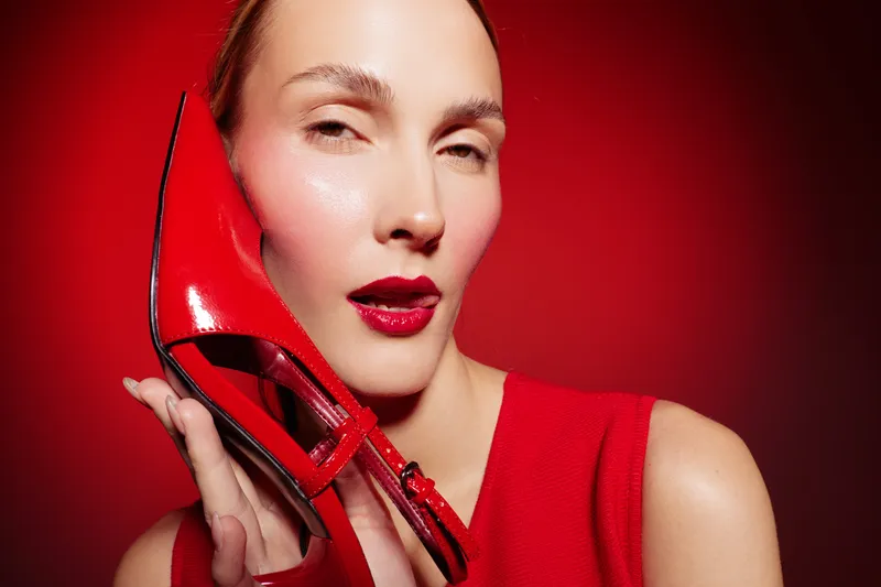 Close-up of a woman with red lipstick holding a shiny red high heel shoe near her face. Red background and red dress create a bold fashion and beauty concept with vibrant colors.