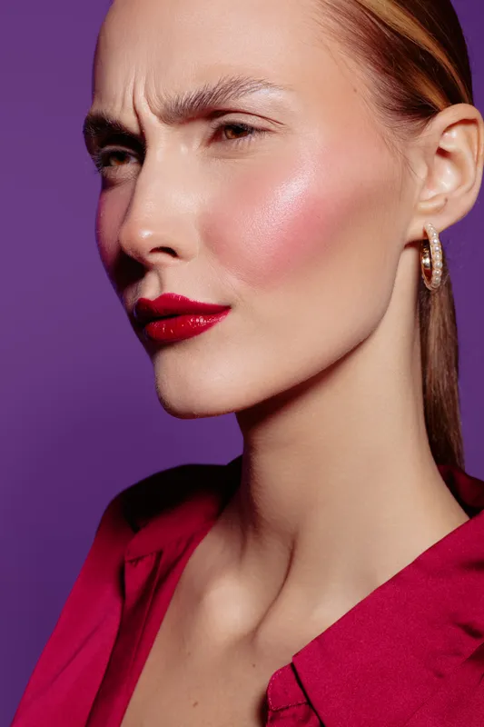Close-up of a woman with red lipstick and shiny pink blush on her cheek, wearing gold hoop earrings and a red blouse. Purple background highlights her serious expression and smooth skin.