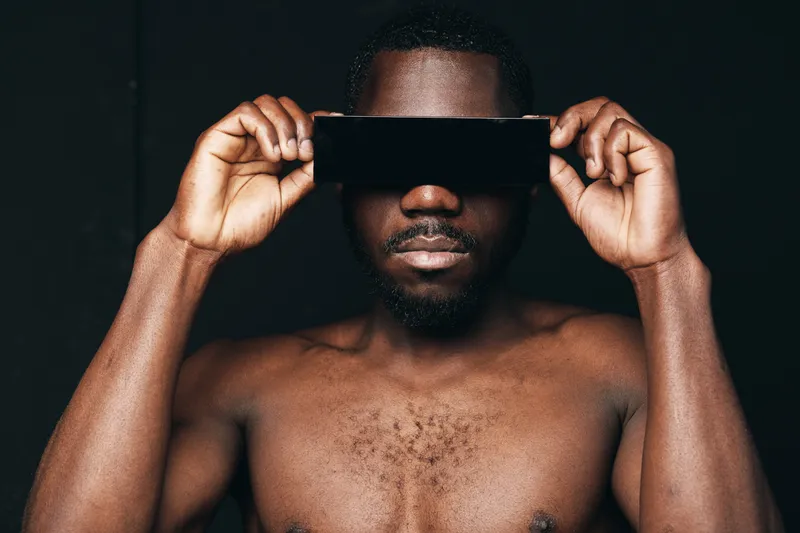 Creative conceptual portrait of African man holding smartphone over eyes with bare chest and dark background in visual style for technology and innovation.