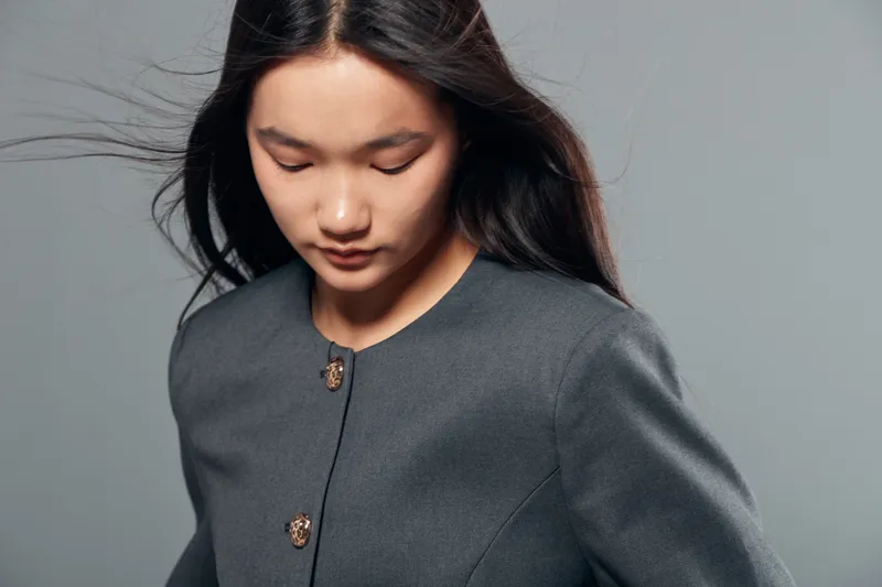 Young woman with long dark hair and closed eyes looking down wearing a gray buttoned jacket against plain gray background in studio portrait.