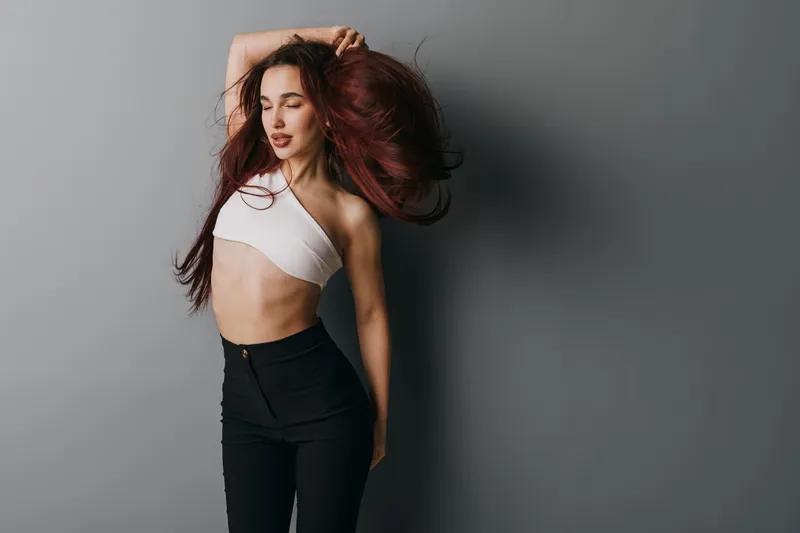 Young woman with long hair in white crop top and black pants posing with eyes closed against gray background. Fashion model showing confident and stylish look indoors.