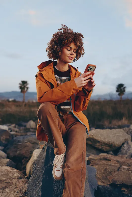 Woman lifestyle outdoors, sitting on rocks and smiling, curly hair and warm jacket, holding a smartphone, sunset light, relaxed confident posture, open landscape background, natural colors