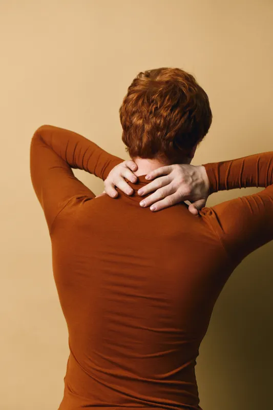 Person with red hair standing with back to camera holding neck with both hands, wearing brown long sleeve shirt against beige background in studio lighting.