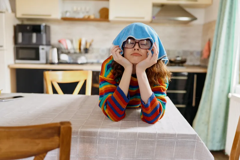 A young girl in a colorful striped sweater rests her chin on her hands, with a blue towel on her head and oversized glasses, posing thoughtfully at a well lit kitchen table