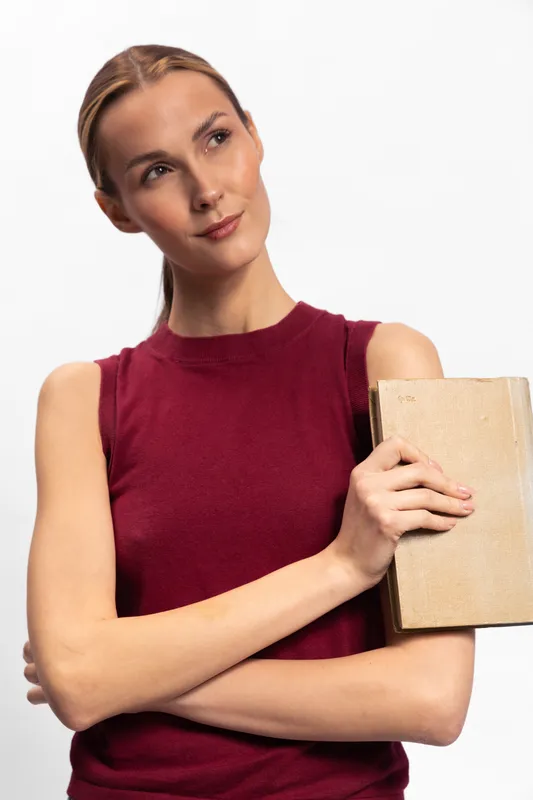 Thoughtful young woman in sleeveless red top holding a book against her chest, looking away on white background. Concept for reading, education, and contemplation.