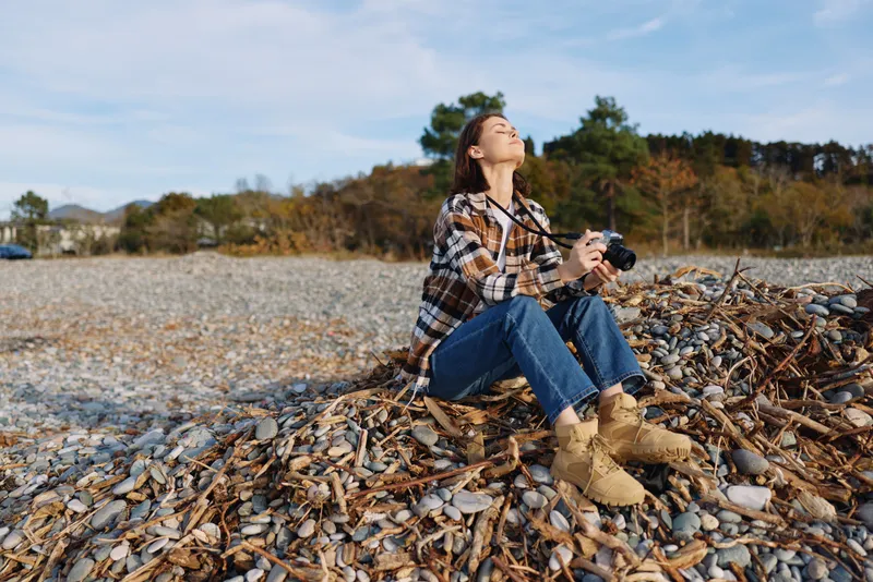 Woman sits on a shell beach, holding a camera, enjoying the outdoors in a casual autumn scene. Scenic coast, pebbles and driftwood, trees in the distance, sunlight on her face, a candid photography