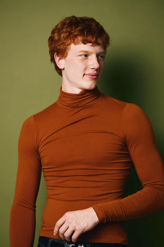 Young man with red hair wearing brown turtleneck sweater standing against green background looking to side with subtle smile and relaxed posture indoors.