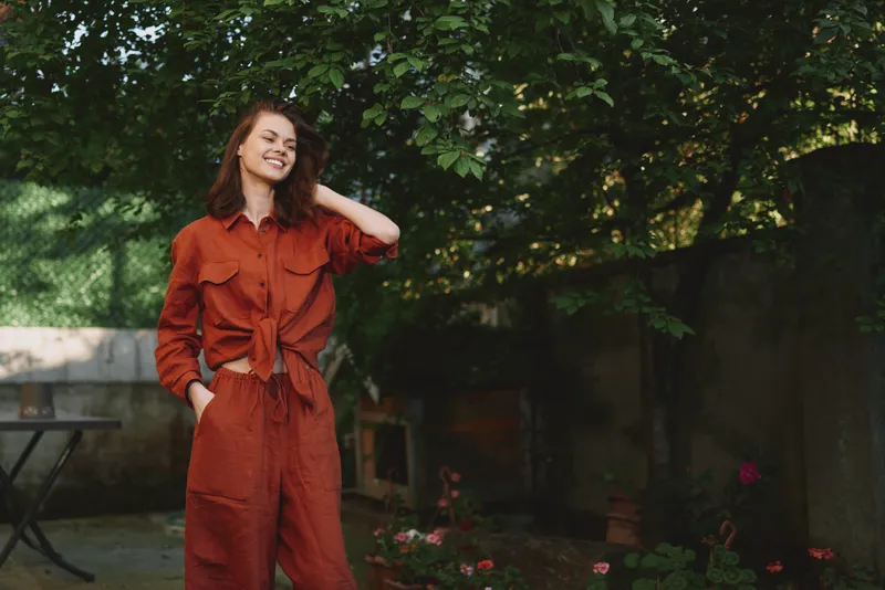 Smiling woman in orange jumpsuit standing in a sunlit garden, casual outdoor lifestyle portrait, natural greenery, relaxed mood, candid moment. Bright energy