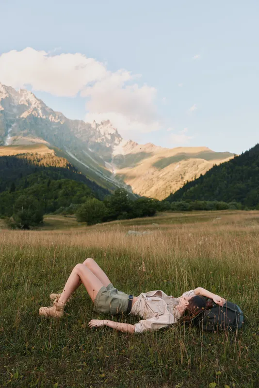 A woman in casual clothes lies on grass in a mountain valley, relaxing with a backpack nearby. The scene shows lifestyle, nature, and outdoor leisure under a clear sky.
