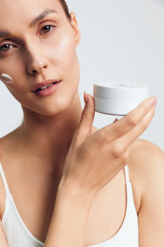 A young woman with clear skin applies cream on her face while holding a white jar in her hand. The bright background emphasizes skin care and beauty routine concepts.