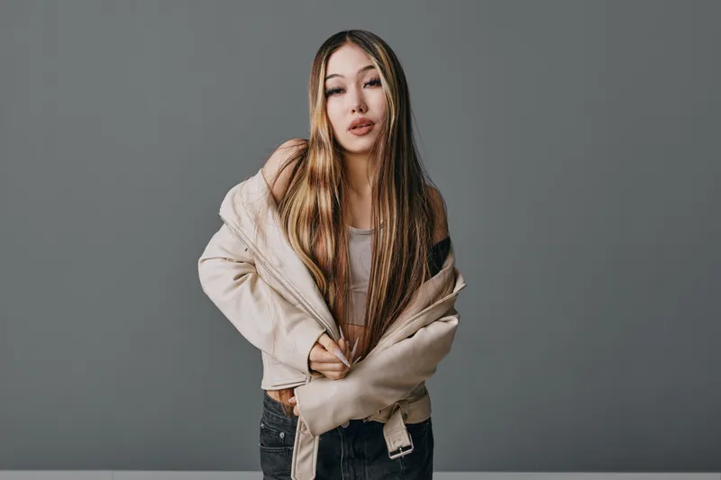 Young woman with long hair wearing casual beige jacket looking confidently at camera on gray background in studio portrait fashion style.