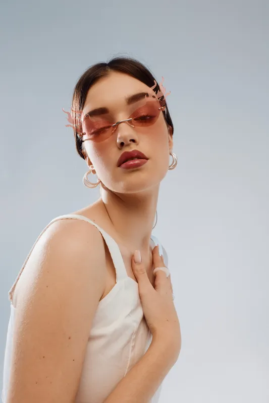 Young woman in pink sunglasses and white top poses with hand on chest against light background. Concept of beauty, fashion, and modern style with confident expression.