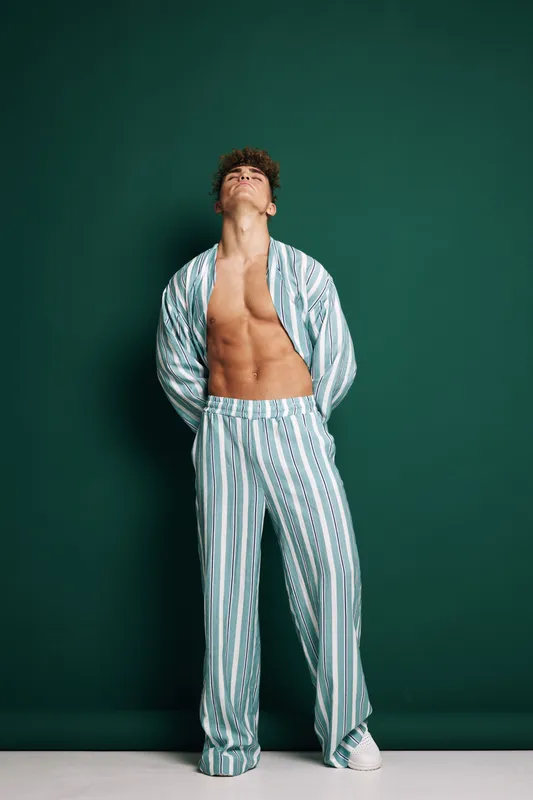 Young man with curly hair wearing striped pajamas standing against green background with hands behind back and head tilted up in relaxed pose.