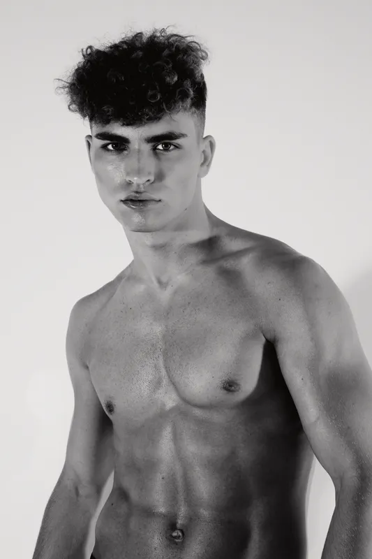 Young man with muscular body and curly hair posing shirtless in black and white studio portrait. Male model with intense look and defined physique standing on neutral background.