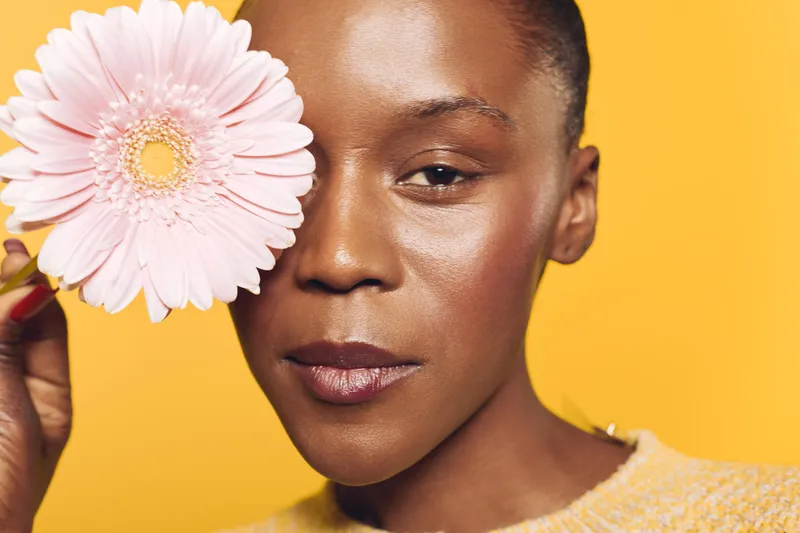 Close-up of a young black woman with dark skin holding a pink flower near her face against a yellow background. Concept of natural beauty, skincare, and floral wellness.