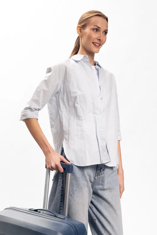 Young woman in casual white shirt and jeans stands with blue suitcase against white background. Concept of travel, trip, vacation, and summer holiday with luggage and relaxed mood.
