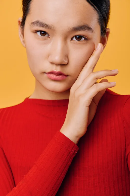 Young woman wearing red sweater posing with hand near face looking confident and calm on yellow background in studio portrait for fashion or beauty concept.