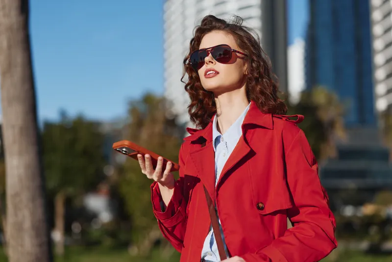 A stylish woman in a red trench coat walks along a sunny city street, holding a smartphone and exuding confidence as modern buildings rise behind her with bold color popping.