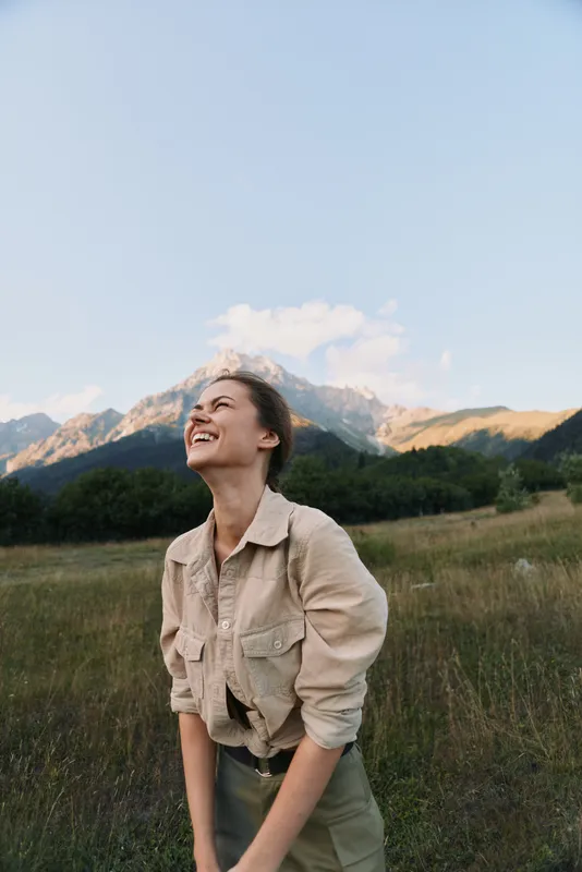 Young woman laughs in a beige shirt in a green meadow with distant mountains and a clear blue sky, enjoying a sunny outdoor moment