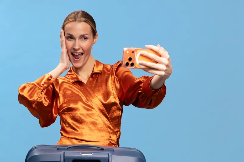Happy young woman in orange satin blouse takes selfie with smartphone while standing behind suitcase on blue background. Concept of travel, vacation, excitement, and modern lifestyle.
