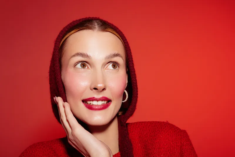Smiling woman with red lips and clear skin wears red hooded sweater, touching face with hand. Bright red background creates vibrant mood. Concept of beauty, skincare, and fashion portrait.