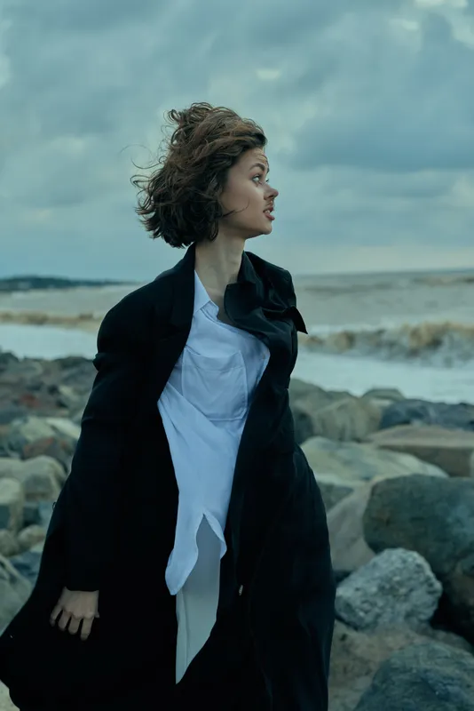 Woman on a windy coastline wearing a dark coat and light shirt, facing the sea with waves and rocks, a dynamic fashion moment at the water edge under a dramatic sky