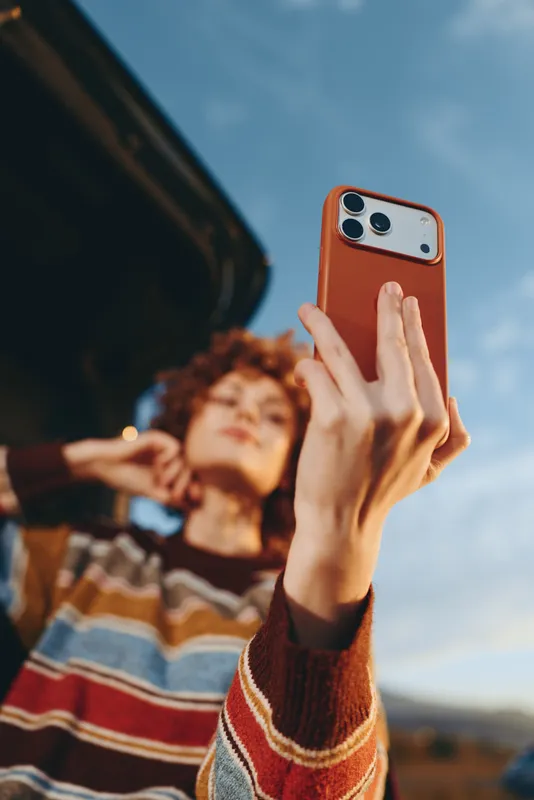 Woman selfie smartphone outdoor lifestyle portrait sky hand sweater travel, holding phone upward against blue sky, blurred friend in background, casual sweater street lifestyle moment today