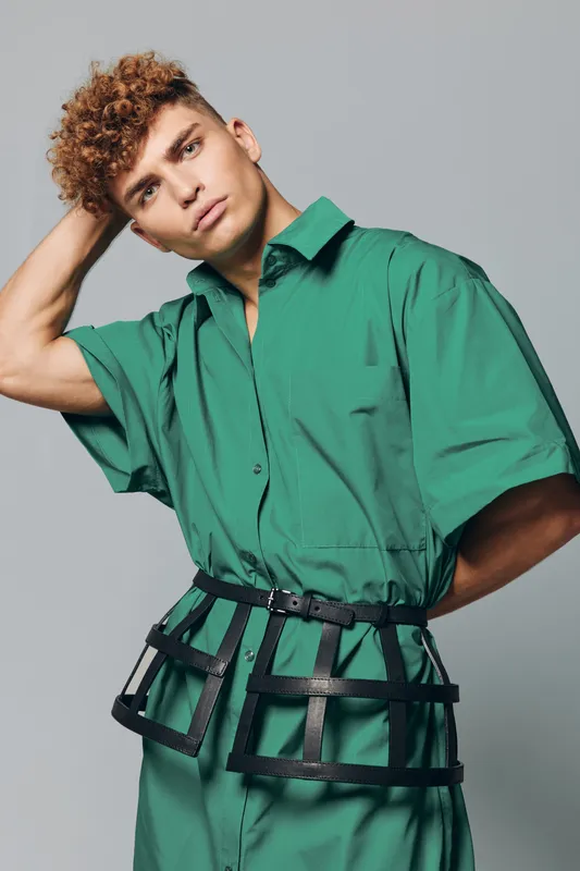 Young man posing in green oversized shirt with large black belt corset standing against grey background. Fashion model with short curly hair and thoughtful expression in studio indoor.