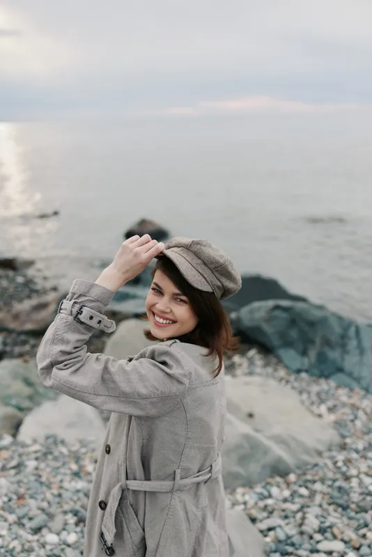 Smiling young woman in a grey coat and hat stands on a rocky beach near calm sea. She looks happy and relaxed, enjoying the cool outdoor atmosphere and natural scenery.