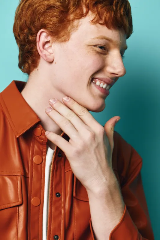 Redhead young man in leather jacket smiling and touching his face on turquoise background. Casual portrait, happy expression and trendy style with natural light and closeup view.