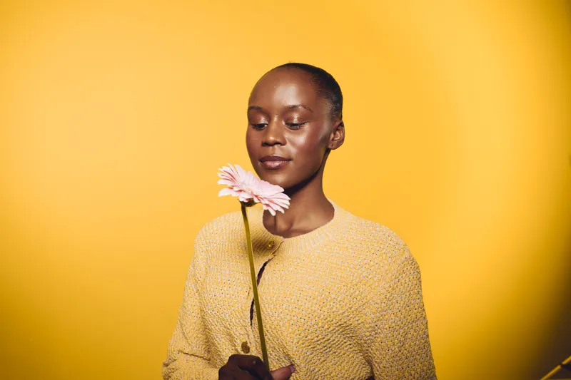 Young black woman with dark skin holding a pink flower in front of yellow background. She wears a yellow sweater and looks down gently. Concept of beauty, calm, and nature.