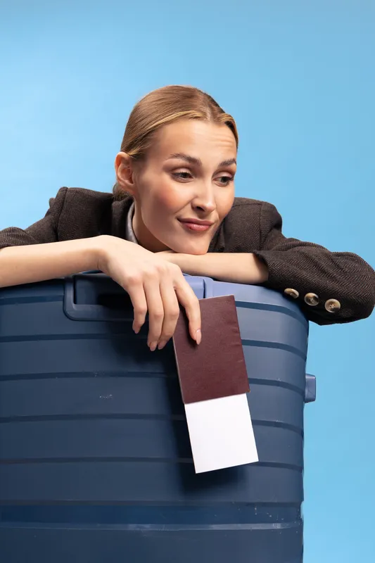 Young woman leans on large blue suitcase holding passport and boarding pass. Travel concept with casual female preparing for trip, isolated on bright blue background.