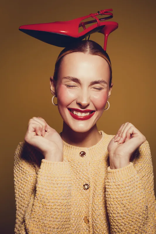 Smiling woman with closed eyes wearing yellow sweater balances red high heel shoe on head. Studio portrait with warm lighting and joyful expression. Concept of fun, fashion, and happiness.
