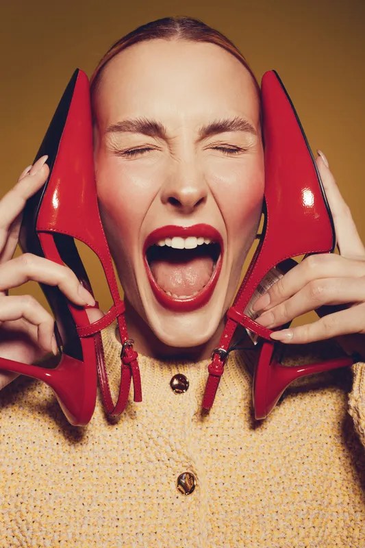 Excited woman with closed eyes holds red high heel shoes near face, wearing yellow sweater. Concept of fashion, style, footwear, and joyful emotions in studio lighting.