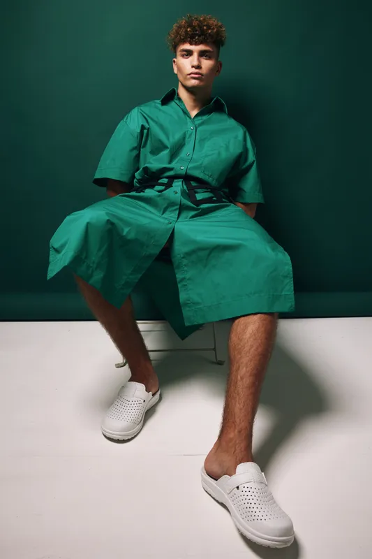 Young man with curly hair wearing a green oversized shirt and matching pants sitting on a white bench with white perforated sneakers on white floor toned with green background studio interior.