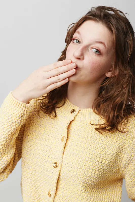 Young woman with curly hair wearing yellow sweater blows a kiss with hand near mouth. Studio portrait on light background. Concept of affection, greeting, and friendly gesture.