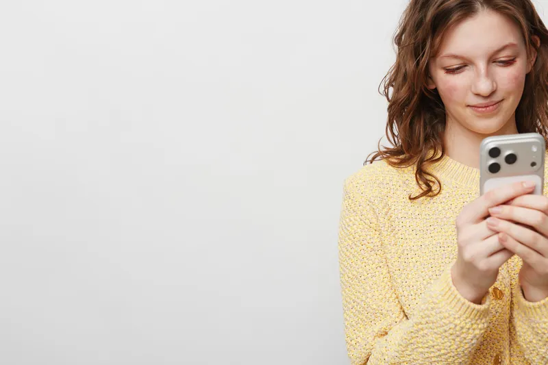 Young woman in yellow sweater looks at smartphone with slight smile, isolated on white background. Concept of technology, communication, social media, and casual lifestyle.