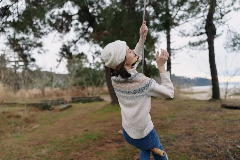 Woman on a swing in a park, wearing a knit sweater and beanie, enjoying a playful moment in a natural outdoor setting